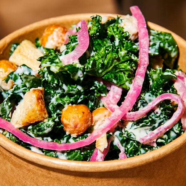 Kale salad with white dressing, pickled red onions, and croutons in a brown paper bowl.