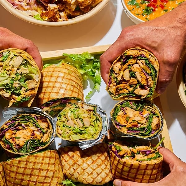 Hands holding chicken wraps with bowls of food and sauce on a wooden tray.