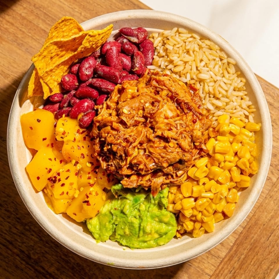 Vibrant bowl with pulled meat, brown rice, kidney beans, corn, guacamole, mango, and tortilla chips.