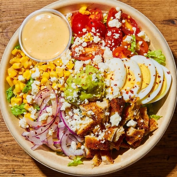 A vibrant salad bowl with chicken, corn, tomatoes, eggs, guacamole, and dressing on a wooden table.