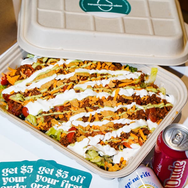 A takeout container of taco salad with sour cream and avocado crema, next to two soda cans and a discount flyer.