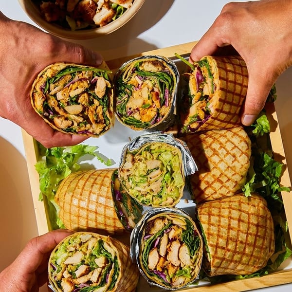 Hands reaching for a tray of grilled wraps, with salads and soup on a white table.